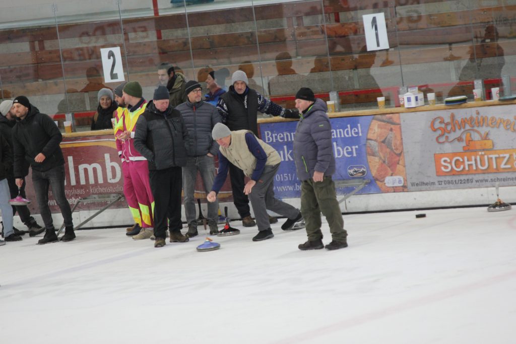 Bild zeigt einen Teilnehmer der gerade den Stock auf der Eisfläche in der Hand hält und mit Schwung gerade losläst. Es sind noch 4 andere Personen zu sehen, die zur Mannschaft gehören und ebenfalls auf dem Eis stehen.
