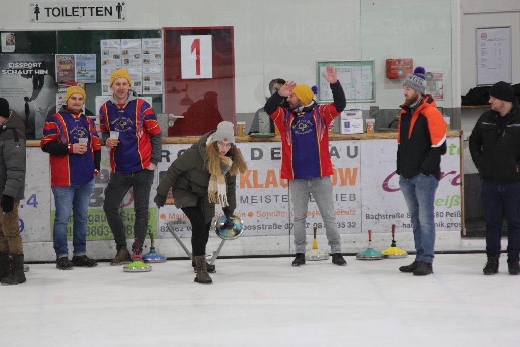Bild zeigt eine Teilnehmerin die gerade den Stock auf der Eisfläche in der Hand hält und mit Schwung gerade losläst. Es sind noch 5 andere Personen zu sehen, die zur Mannschaft gehören und ebenfalls auf dem Eis stehen.