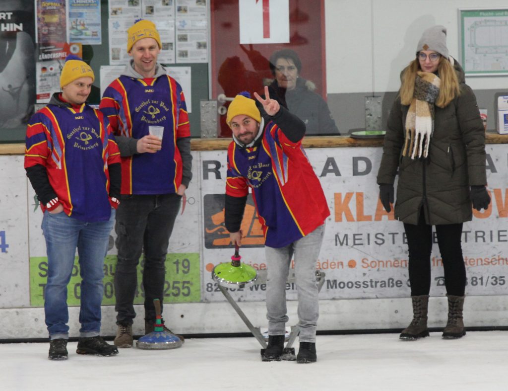 Bild zeigt einen Teilnehmer der gerade den Stock auf der Eisfläche in der Hand hält und mit Schwung gerade losläst. Es sind noch drei andere Personen zu sehen, die zur Mannschaft gehören und ebenfalls auf dem Eis stehen.