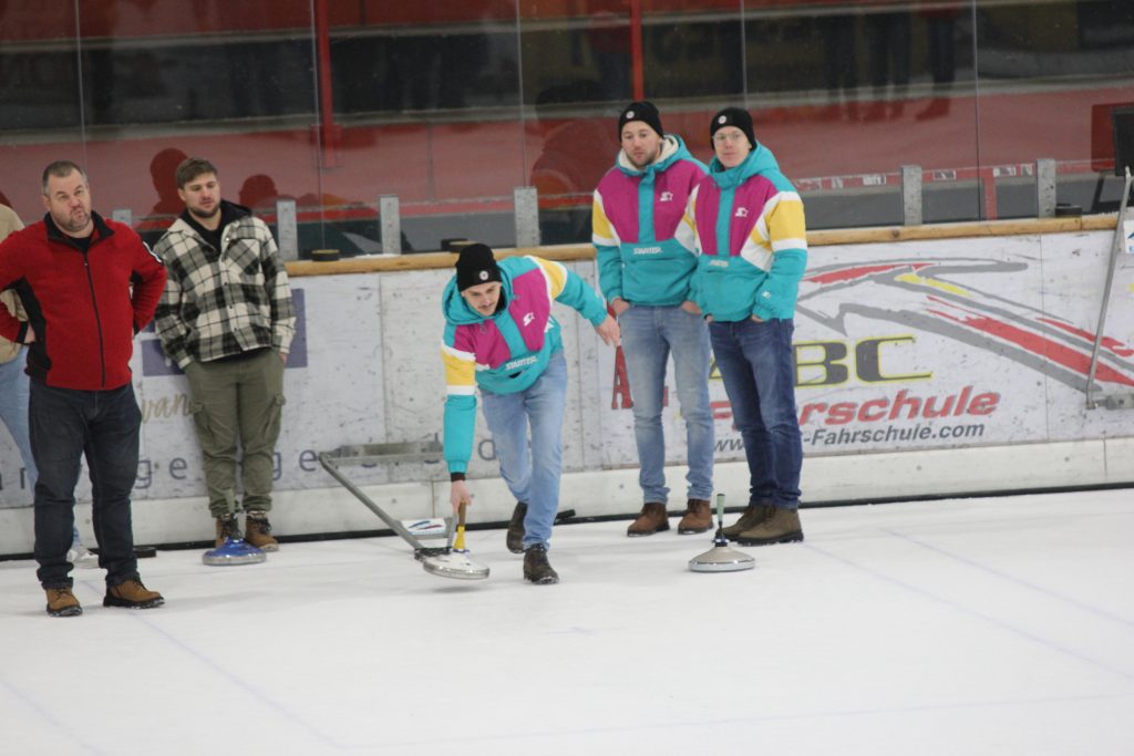 Bild zeigt einen Teilnehmer der gerade den Stock auf der Eisfläche in der Hand hält und mit Schwung gerade losläst. Es sind noch 4 andere Personen zu sehen, die zur Mannschaft gehören und ebenfalls auf dem Eis stehen.