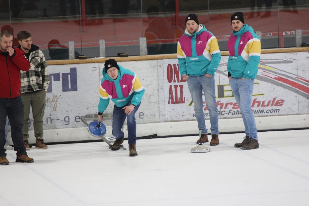 Bild zeigt einen Teilnehmer der gerade den Stock auf der Eisfläche in der Hand hält und mit Schwung gerade losläst. Es sind noch 4 andere Personen zu sehen, die zur Mannschaft gehören und ebenfalls auf dem Eis stehen.