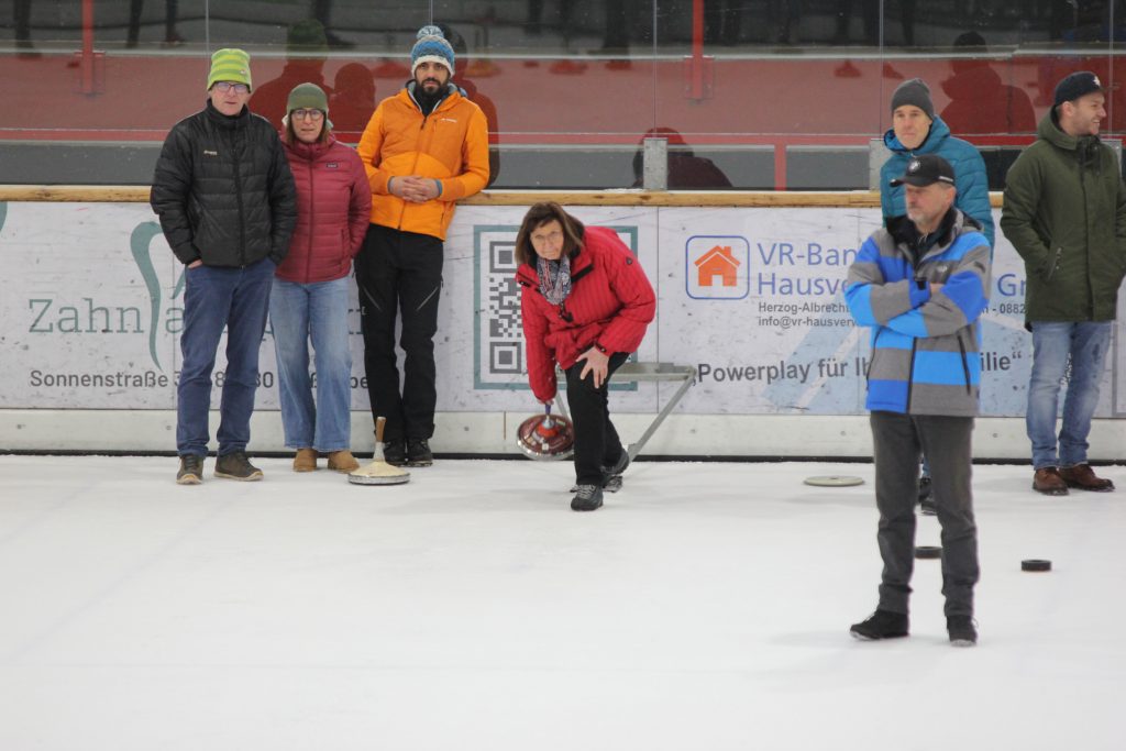 Bild zeigt eine Teilnehmerin die gerade den Stock auf der Eisfläche in der Hand hält und mit Schwung gerade losläst. Es sind noch 5 andere Personen zu sehen, die zur Mannschaft gehören und ebenfalls auf dem Eis stehen.