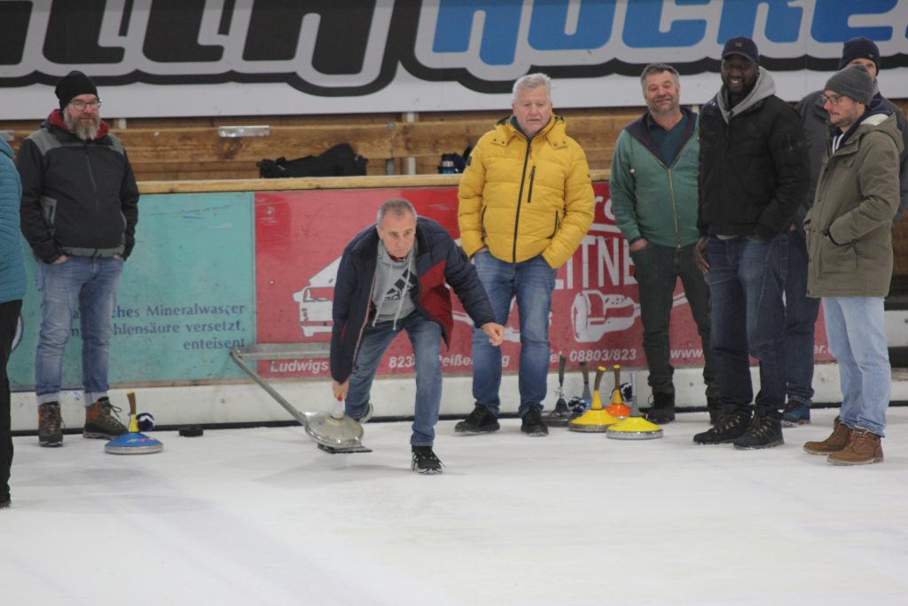 Bild zeigt einen Teilnehmer der gerade den Stock auf der Eisfläche in der Hand hält und mit Schwung gerade losläst. Es sind noch 5 andere Personen zu sehen, die zur Mannschaft gehören und ebenfalls auf dem Eis stehen.