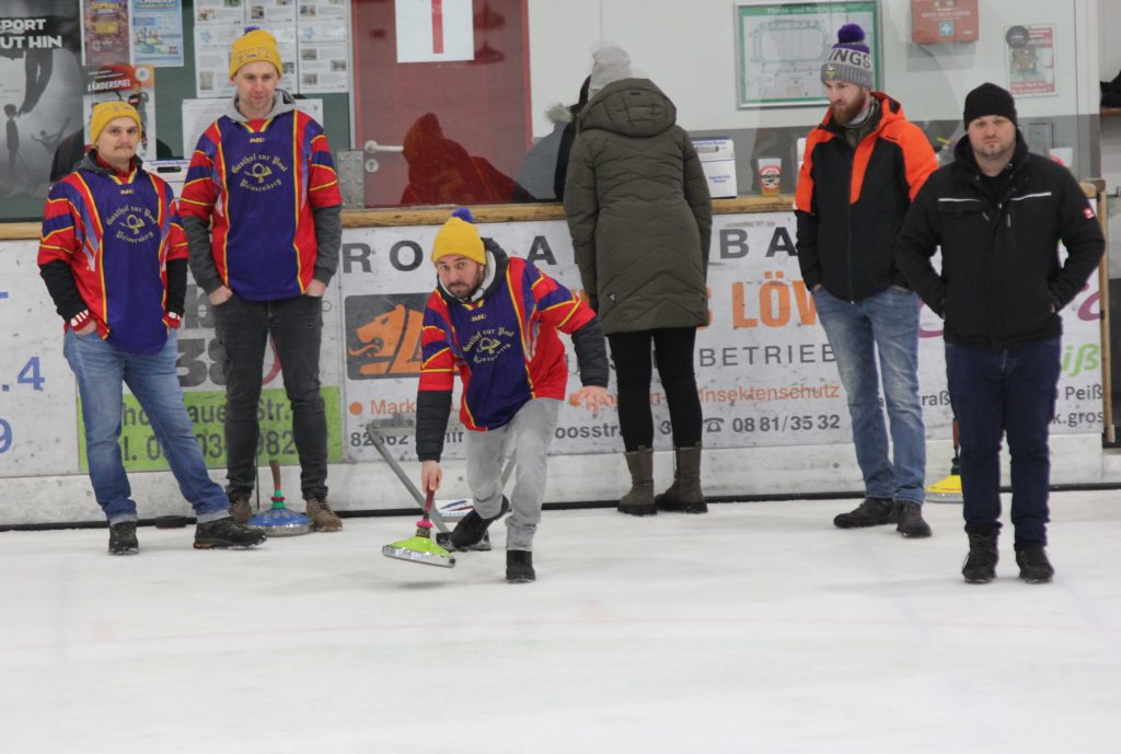 Bild zeigt einen Teilnehmer der gerade den Stock auf der Eisfläche in der Hand hält und mit Schwung gerade losläst. Es sind noch 4 andere Personen zu sehen, die zur Mannschaft gehören und ebenfalls auf dem Eis stehen.