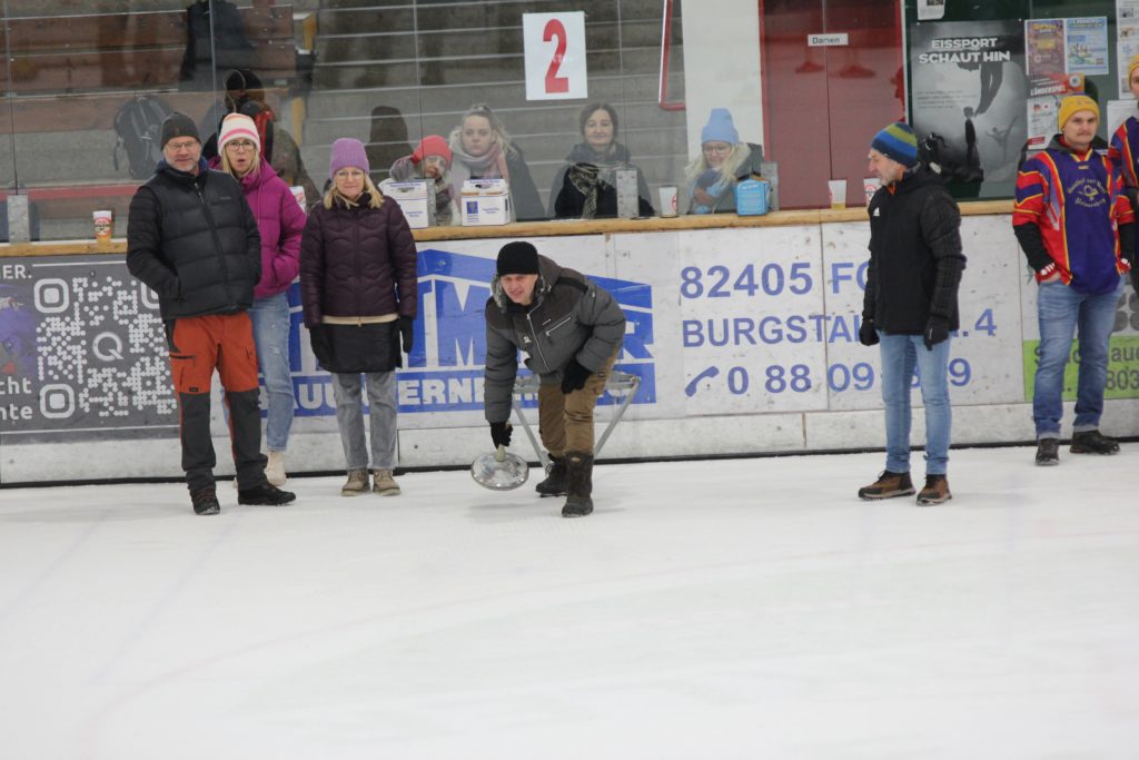 Bild zeigt einen Teilnehmer der gerade den Stock auf der Eisfläche in der Hand hält und mit Schwung gerade losläst. Es sind noch 4 andere Personen zu sehen, die zur Mannschaft gehören und ebenfalls auf dem Eis stehen.