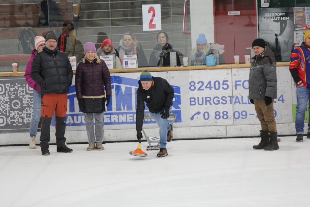 Bild zeigt einen Teilnehmer der gerade den Stock auf der Eisfläche in der Hand hält und mit Schwung gerade losläst. Es sind noch 4 andere Personen zu sehen, die zur Mannschaft gehören und ebenfalls auf dem Eis stehen.